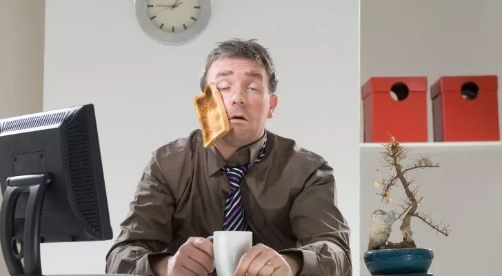 A man looking tired holding a coffee with a slice of toast stuck to his cheek