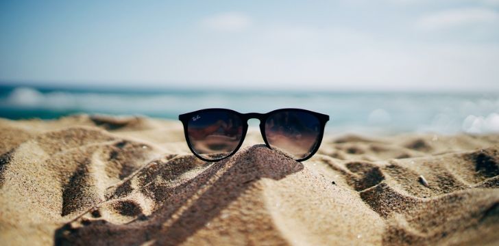 A pair of Ray Ban sunglasses on sand at the beach with the ocean behind