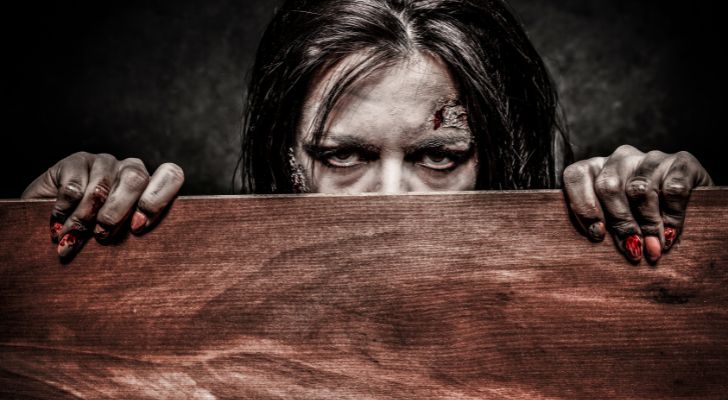 A female zombie peering over the top of a plank of wood