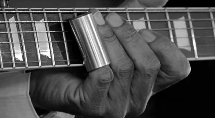 A hand playing a guitar with a metal cylinder covering one finger