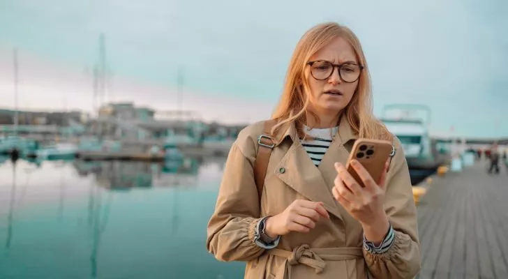 A woman standing on a dock looks at her phone in a confused manner