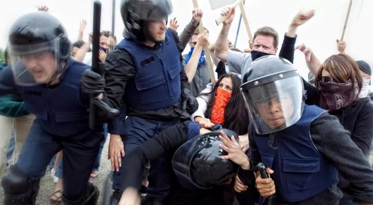 An angry crowd and police in riot gear have a physical confrontation