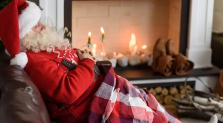 Santa sleeps in a chair in front of a fireplace while covered in a blanket