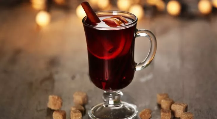 A glass of mulled wine with a stick of cinnamon sticking out surrounded by brown sugar cubes