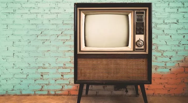 An old TV set with legs stands against a plain brick wall