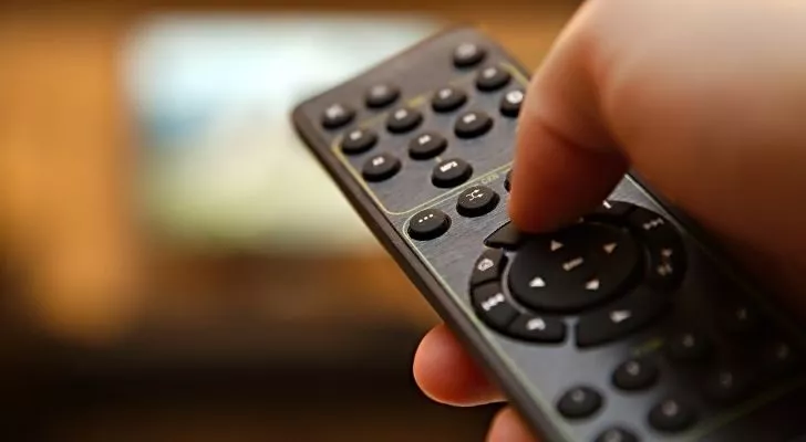 A hand holds a TV remote and presses a button while pointing it at a television