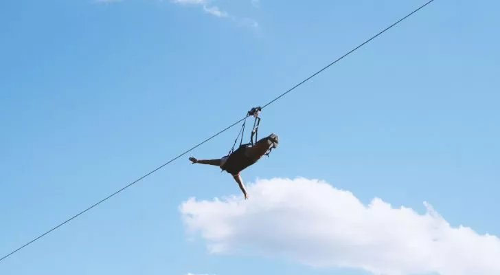 Someone rides a zipline against a pale blue sky with a cloud