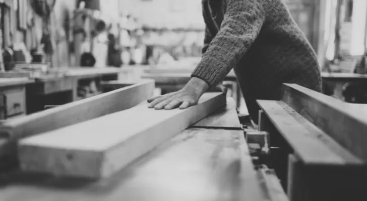 A man in a woodworking shop leans on a thick plank of wood