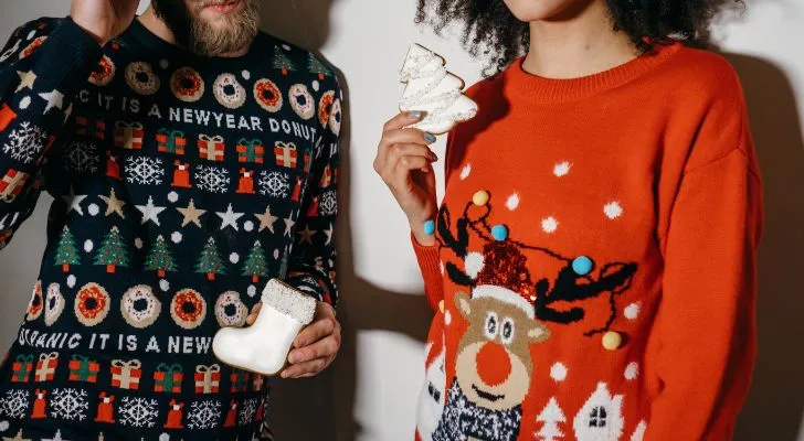 Two people holding Christmas cookies while wearing bright, heavily decorated Christmas sweaters