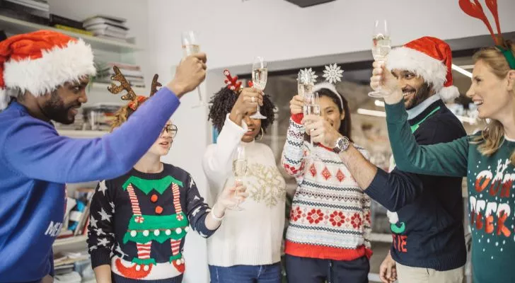 A group of friends wearing overly ornate Christmas sweaters toast with champagne