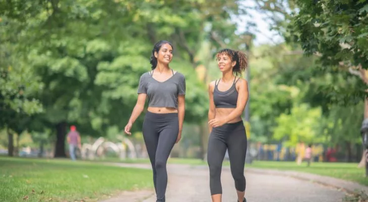 Two women in exercise clothing walk through a park
