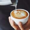 Close-up of a person holding a cup of coffee with intricate latte art being poured from a metal pitcher