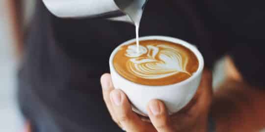 Close-up of a person holding a cup of coffee with intricate latte art being poured from a metal pitcher