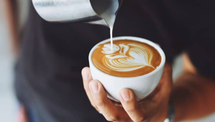 Close-up of a person holding a cup of coffee with intricate latte art being poured from a metal pitcher