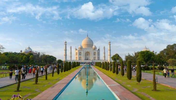People walking along a pathway leading to the Taj Mahal, an iconic white marble mausoleum, surrounded by gardens and reflecting pools under a blue sky