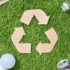 Recycling symbol made of cardboard surrounded by a paper cup, plastic bottle, glass bottle, light bulb, and aluminum can on grass