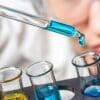 Close-up of a person in safety goggles using a pipette to add blue liquid to test tubes, likely related to significant chemical reactions