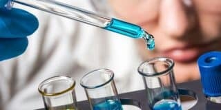 Close-up of a person in safety goggles using a pipette to add blue liquid to test tubes, likely related to significant chemical reactions