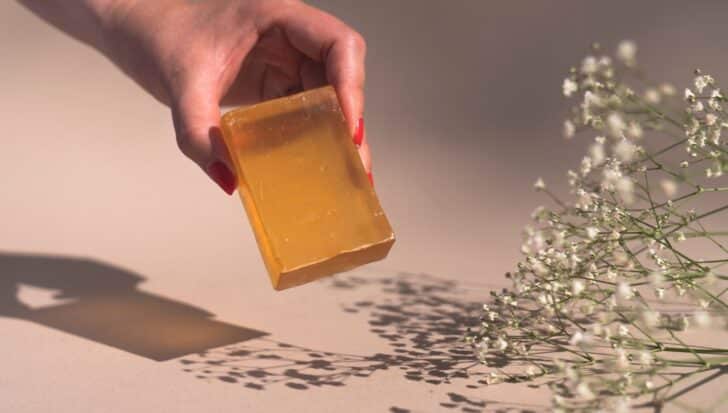 A hand holding a translucent bar of soap next to delicate white flowers on a light background