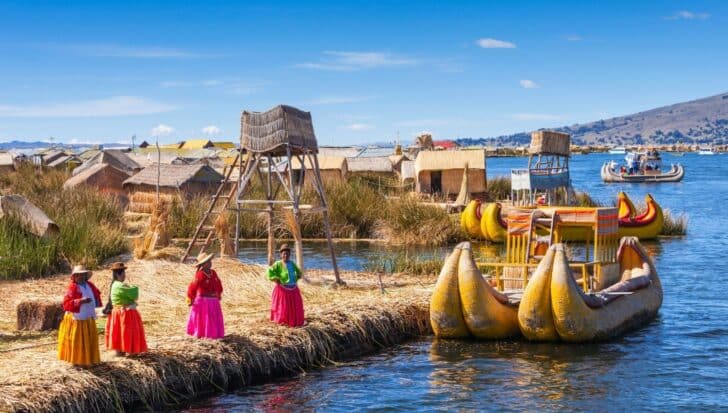 Scenic view of Lake Titicaca with people in traditional clothing standing on a reed island, colorful boats, and huts in the background