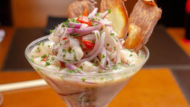 A glass filled with ceviche, Peru's national dish, featuring raw fish marinated in citrus juices, garnished with red onions, chili, and herbs, with potato slices on the side