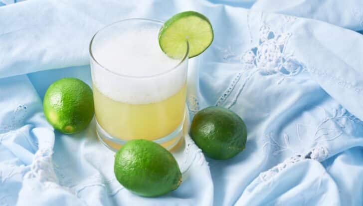 A glass of Peru's national drink garnished with a lime slice, surrounded by whole limes on a blue embroidered fabric