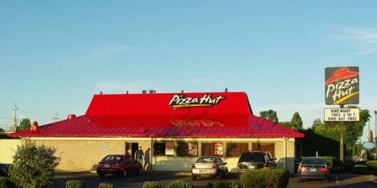Exterior of a Pizza Hut restaurant with a red roof and several cars parked outside, displaying a sign advertising Kids Night
