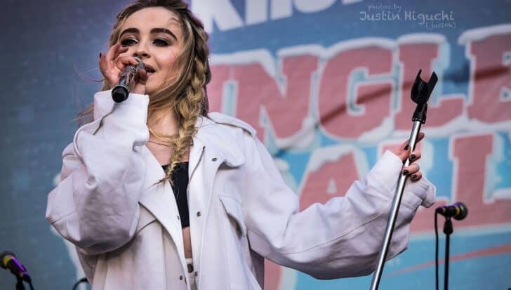 Sabrina Carpenter performing on stage, holding a microphone, wearing a white jacket with a colorful backdrop behind her
