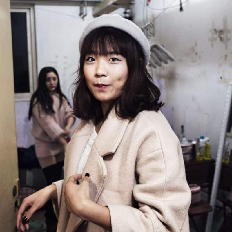 Two people inside a small, cluttered living space with worn walls and household items