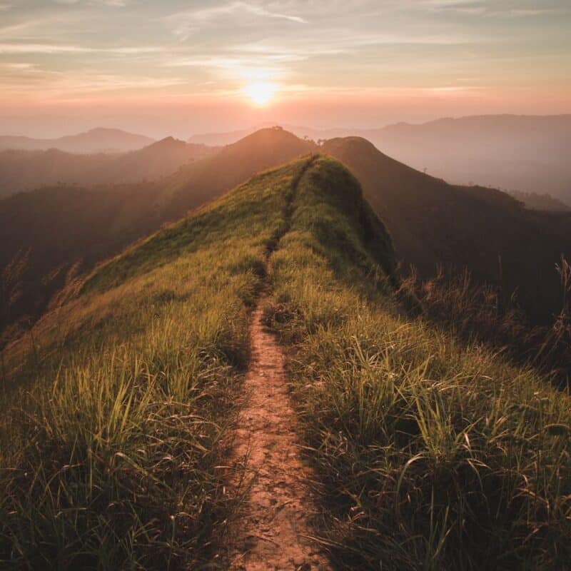 A narrow dirt path on a grassy mountain ridge leading towards the sunrise, surrounded by rolling hills