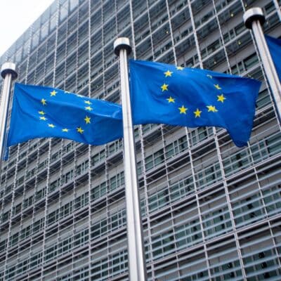 Three European Union flags with twelve stars each in front of a modern building