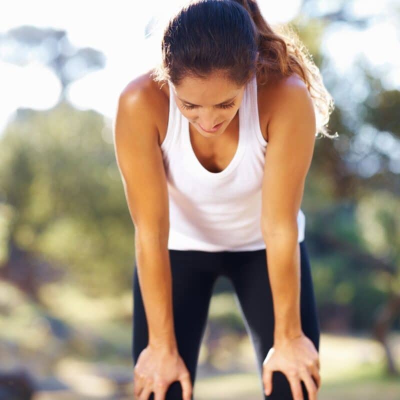 A woman in a white tank top and black leggings bending forward outdoors