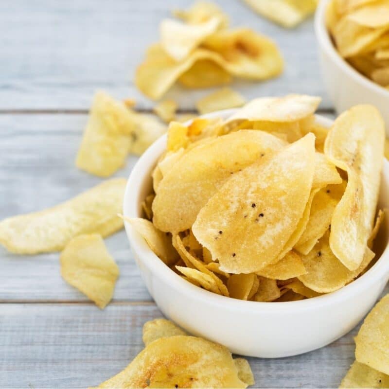 Bowl of crispy potato chips on a wooden surface with scattered chips around