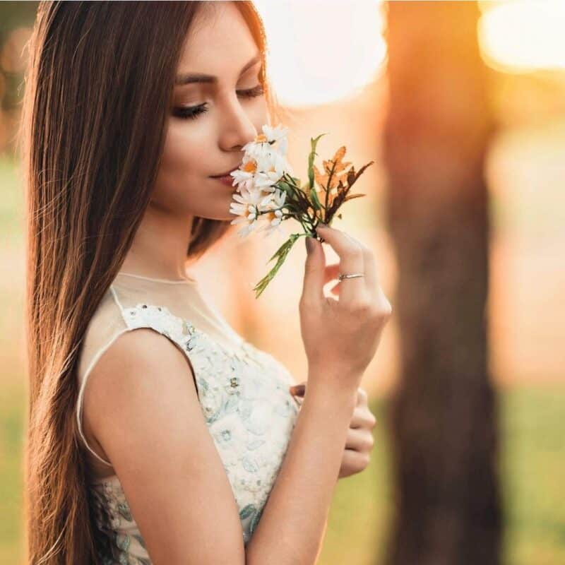 A young woman with long hair smelling a bouquet of white flowers in a sunlit outdoor setting