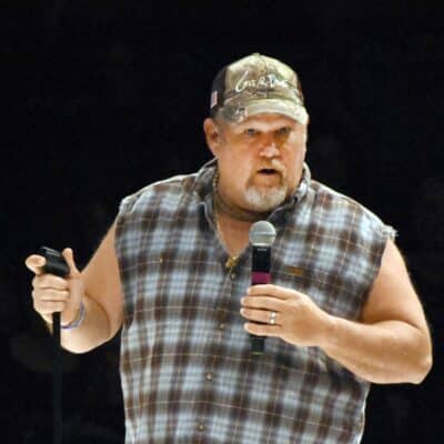 Daniel Lawrence Whitney, known as Larry the Cable Guy, performing on stage holding a microphone and wearing a sleeveless plaid shirt and a cap.