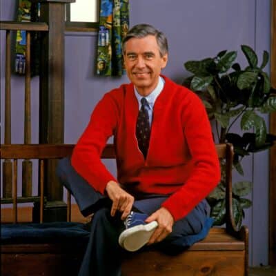 Mister Rogers is sitting on a wooden bench, wearing a hand-knitted red sweater