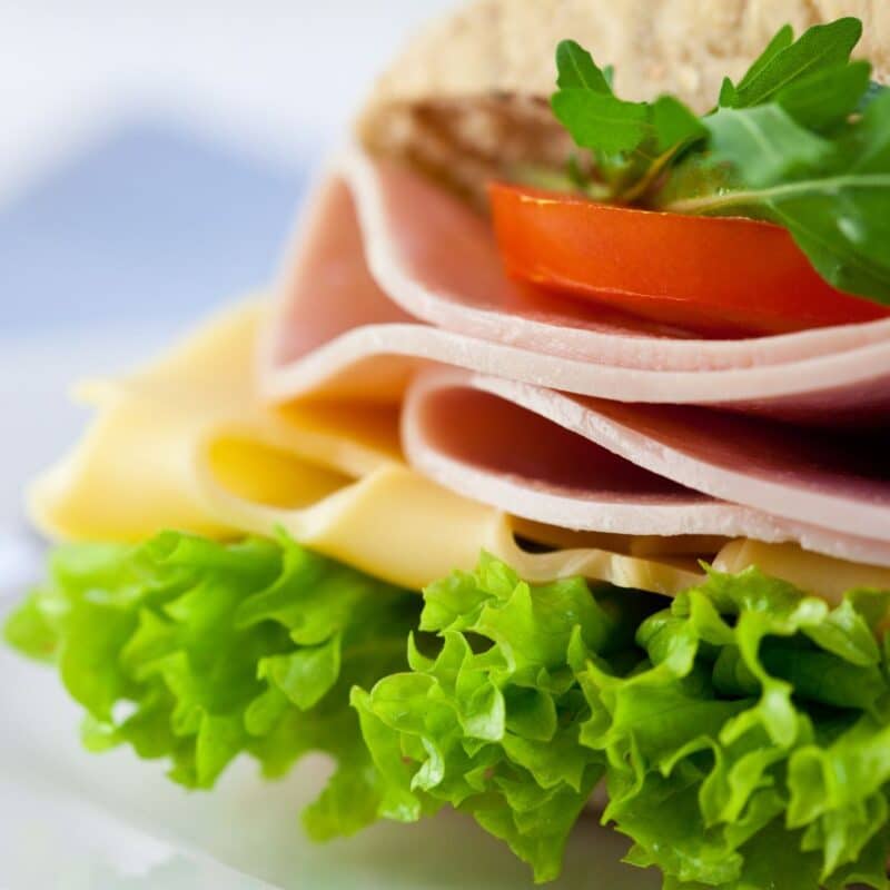 Close-up of a sandwich with layers of cooked meat, cheese, lettuce, tomato, and leafy greens