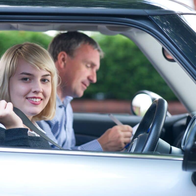 A young person in a car putting on a seatbelt with an adult sitting next to them holding a clipboard