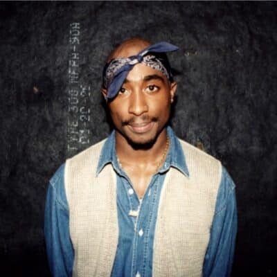 Tupac wearing a denim shirt and beige vest with a blue bandana on his head, standing against a dark textured background