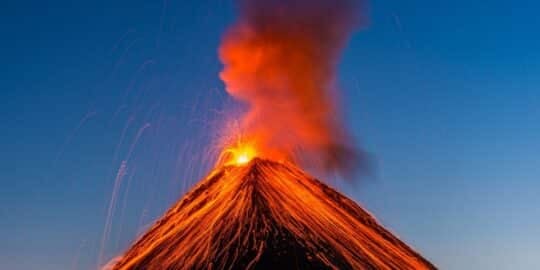 A volcano erupting with bright lava and smoke against a clear blue sky