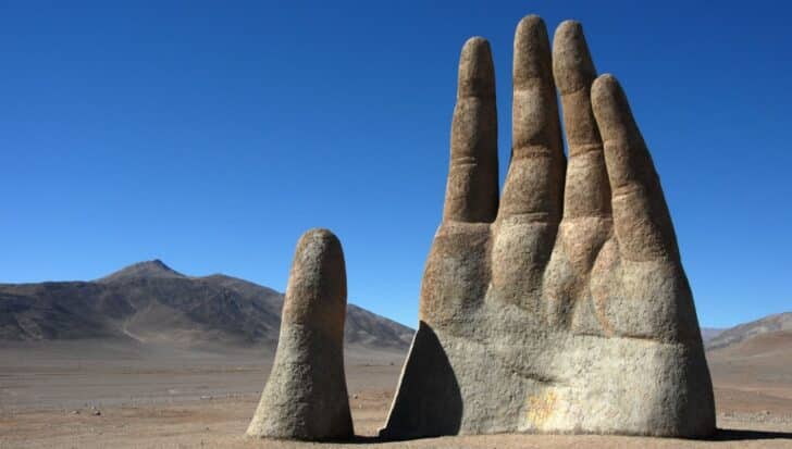 La Mano del Desierto, a large concrete sculpture of a hand emerging from the desert landscape, with mountains in the background under a clear blue sky in Chile