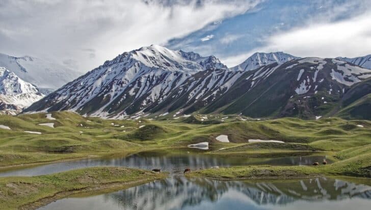 Kyrgyzstan's mountainous landscape with snow-capped peaks and grassy hills surrounding a reflective lake at high elevation