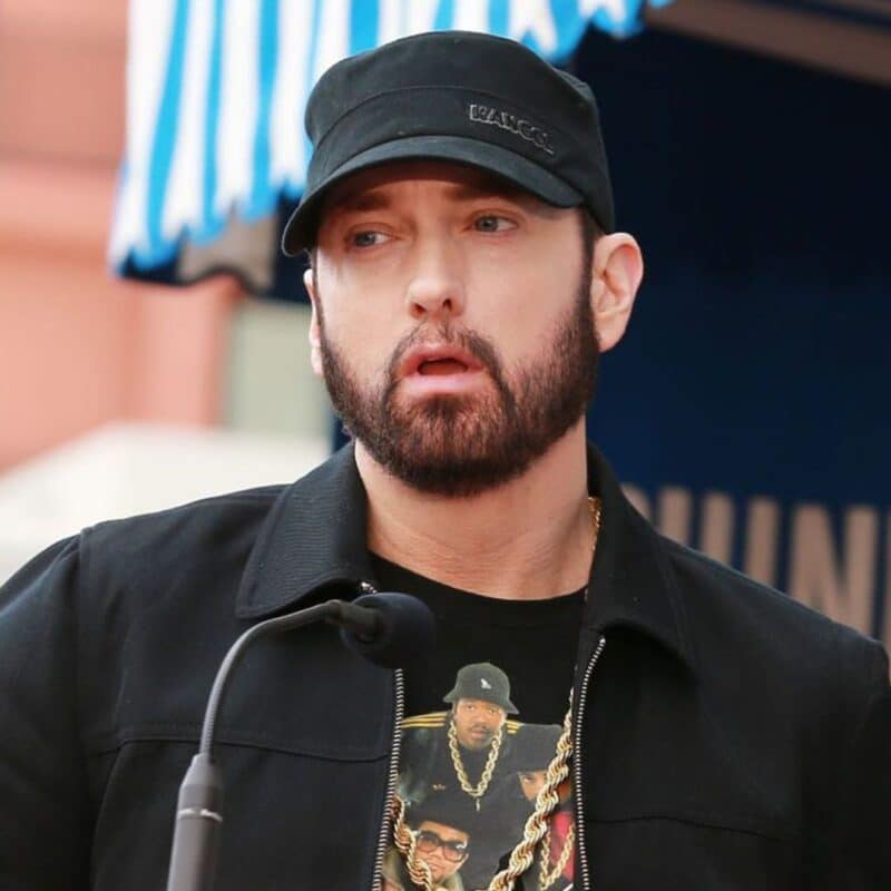 Eminem wearing a black cap and jacket, standing behind a microphone