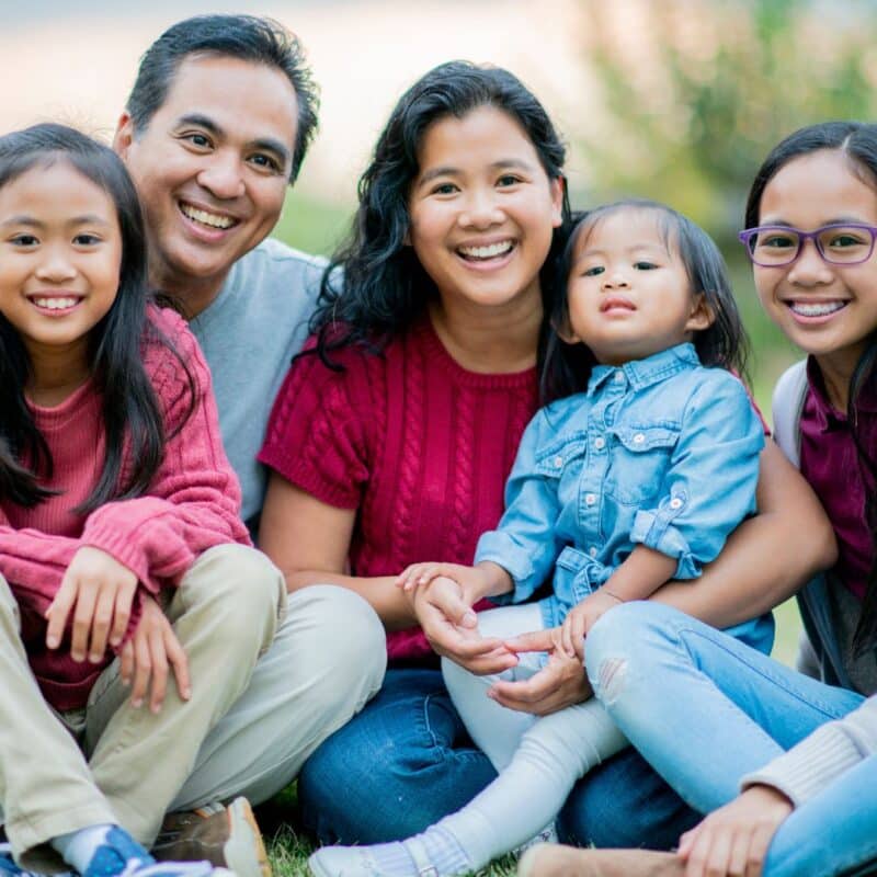 Happy family of five sitting together outdoors, including two adults and three children smiling at the camera