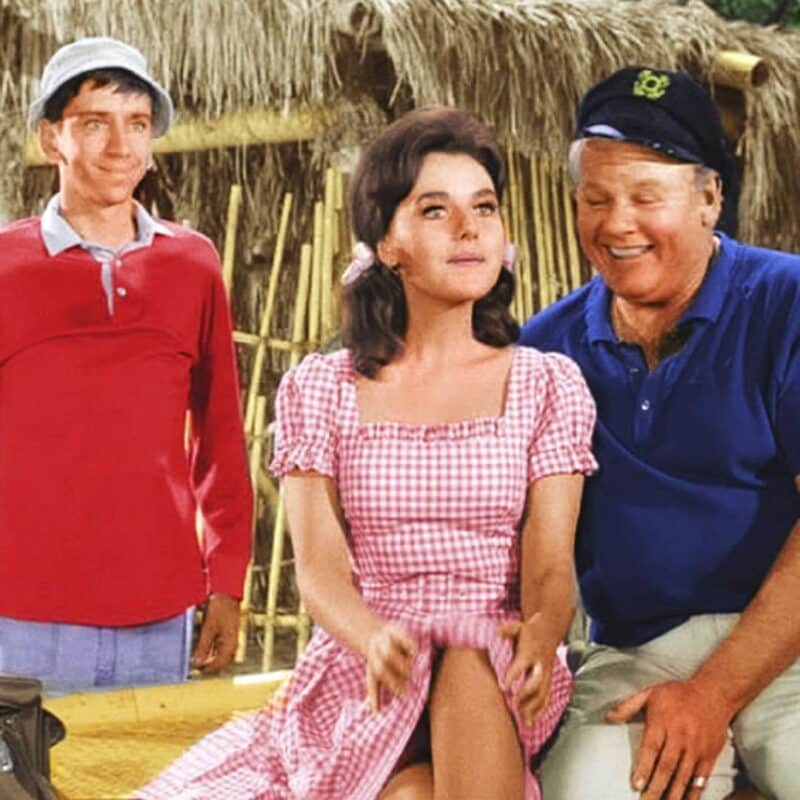 Gilligan, Mary Ann, and the Skipper from Gilligan's Island standing in front of a bamboo hut
