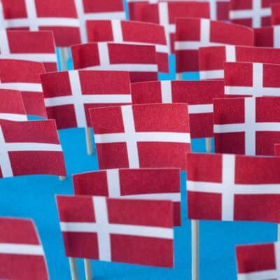 Many small Danish flags with white crosses on a red background displayed on sticks against a blue backdrop