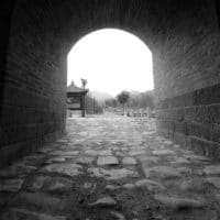 View through a stone tunnel entrance opening to a paved path with trees and a traditional building, with mountains in the background
