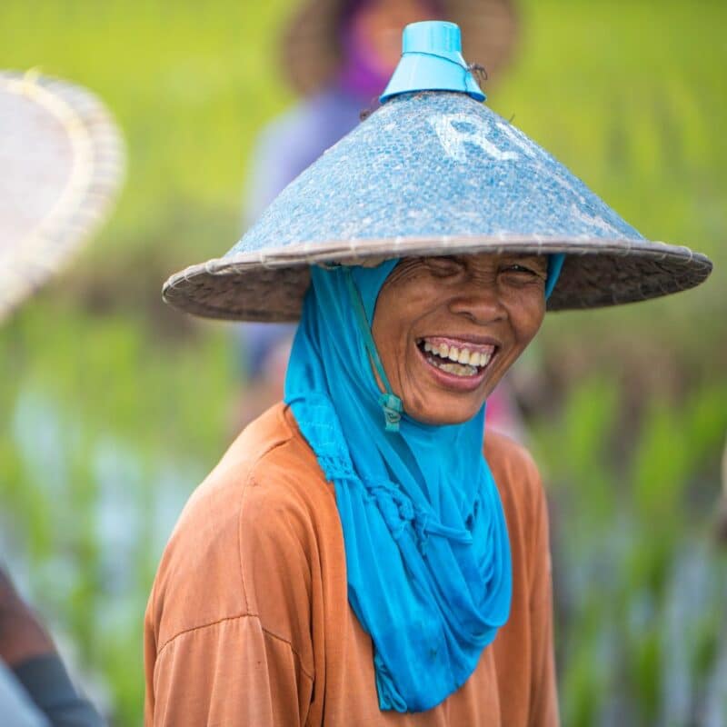 A person wearing a blue headscarf and a conical hat smiles broadly in a green outdoor setting