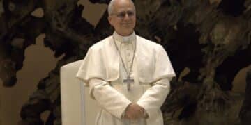 Pope Leo XIV, standing with hands folded, against a dark textured background