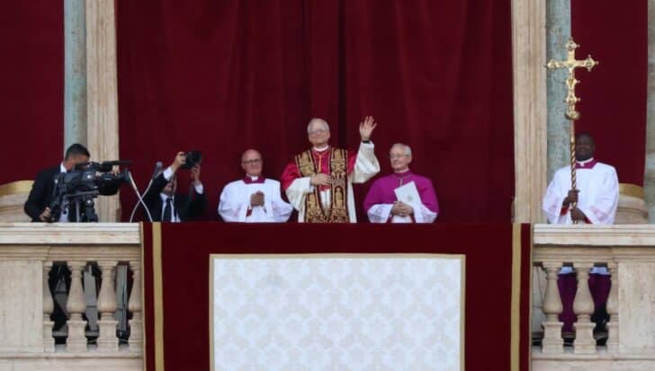 Pope Leo XIV waves from a balcony alongside clergy members, with photographers capturing the moment